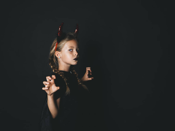 little girl in halloween devil costume with horns on head looking at camera with frightening gesture on black background  - Fotografie, Obrázek