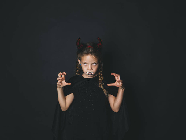 little girl in halloween devil costume with horns on head and white artificial fangs in mouth looking at camera with frightening gesture on black background  - Foto, afbeelding