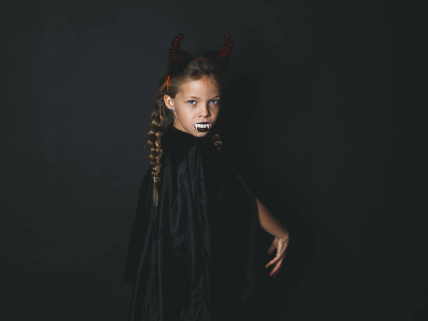 little girl in halloween devil costume with horns on head and black cloak looking at camera while showing white artificial fangs in mouth on black background  - Fotografie, Obrázek