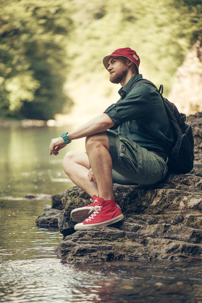 Tourist guy with backpack sitting near lake in a wild summer wood. Expedition - Foto, Bild