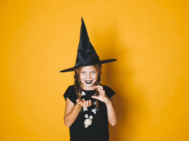 little girl in halloween witch costume and black hat looking at camera with frightening gesture on orange background   - Photo, Image