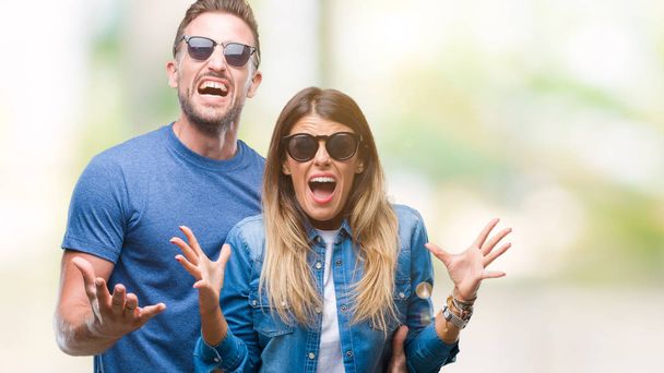 Young couple in love wearing sunglasses over isolated background crazy and mad shouting and yelling with aggressive expression and arms raised. Frustration concept. - 写真・画像