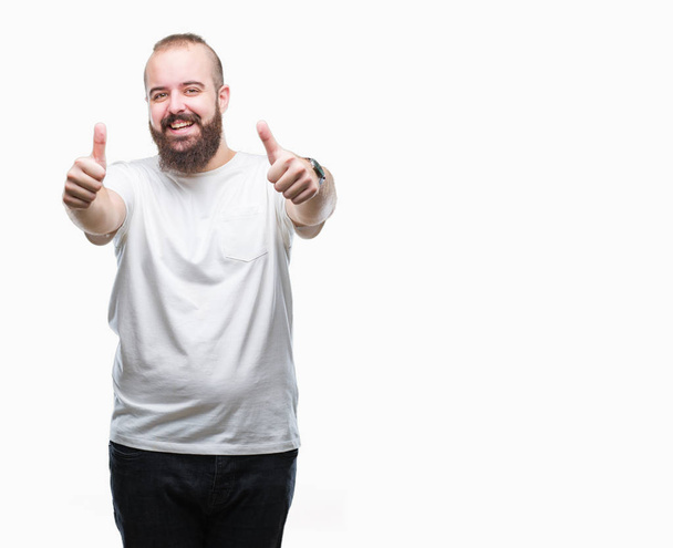 Jeune homme hipster caucasien portant t-shirt décontracté sur fond isolé approuvant faire geste positif avec la main, pouces levés souriant et heureux pour le succès. En regardant la caméra, geste gagnant
. - Photo, image