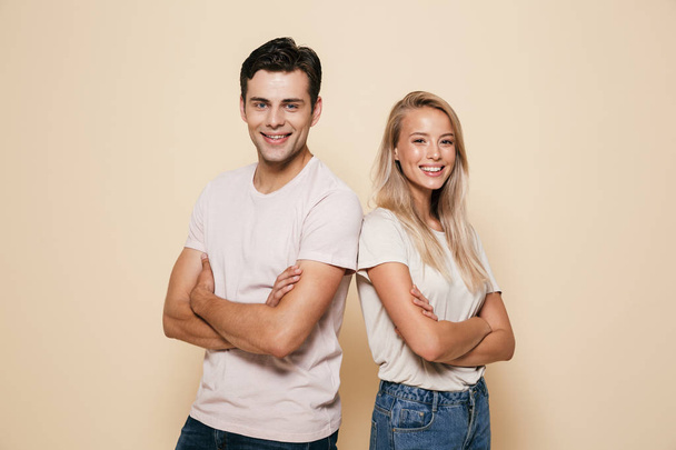 Portrait of a smiling young couple standing together over beige background, arms folded - Фото, зображення