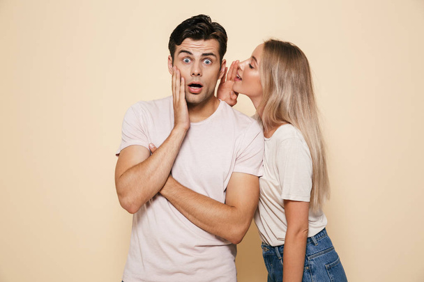 Portrait of a shocked young couple standing together over beige background, telling secrets - Foto, afbeelding