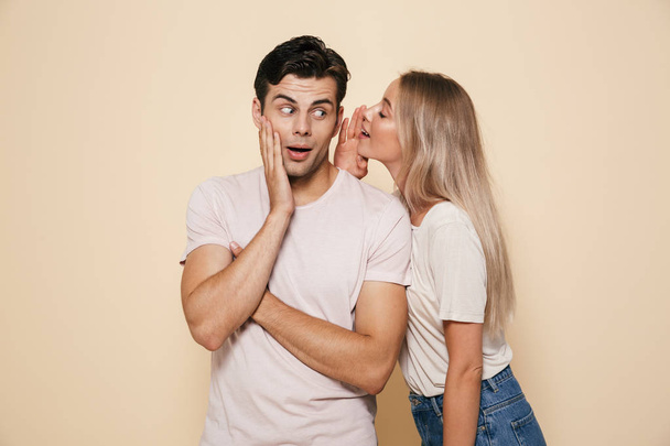 Portrait of a surprised young couple standing together over beige background, telling secrets - Foto, Imagem