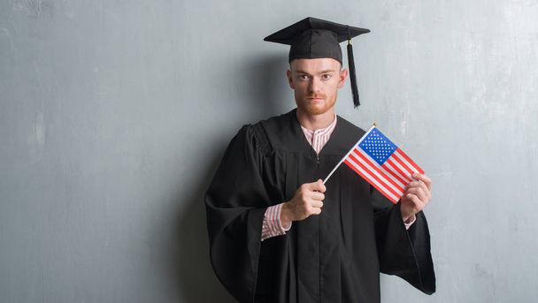 Jeune homme rousse sur le mur gris grunge portant uniforme diplômé tenant drapeau américain avec une expression confiante sur la pensée intelligente visage sérieux
 - Photo, image
