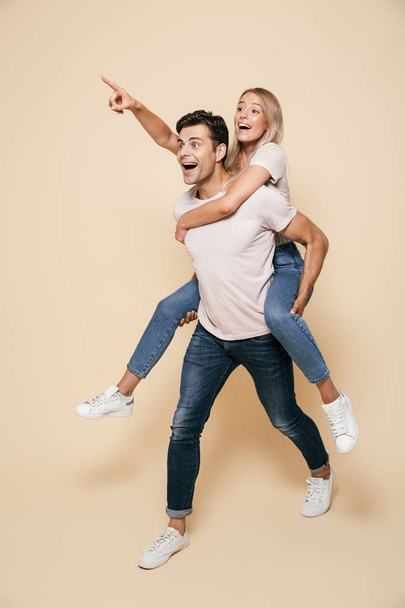Full length portrait of a cheerful young couple standing together over beige background, piggyback ride, pointing away - Foto, afbeelding
