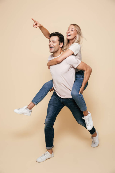 Full length portrait of a cheerful young couple standing together over beige background, piggyback ride, pointing finger away - Фото, зображення