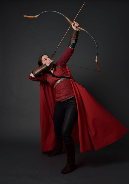 retrato de longitud completa de chica morena con traje medieval rojo y capa, sosteniendo un arco y flecha. pose de pie sobre fondo gris estudio
. - Foto, imagen