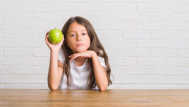 スマートの顔は深刻な思考に自信を持って式に新鮮な青リンゴを食べるテーブルの上に座って若いヒスパニックの子供 - 写真・画像