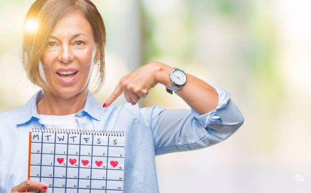 Старша іспаномовна жінка, що тримає календар менструацій на ізольованому фоні з несподіваним обличчям, спрямованим пальцем до себе
 - Фото, зображення