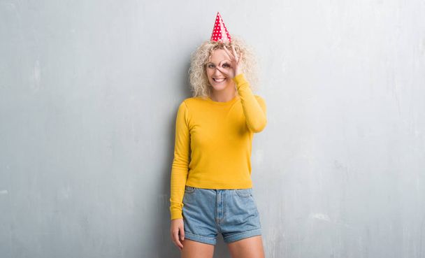 Fiatal szőke nő fölött grunge szürke fal rajta születésnapi kalapot csinál rendben gesztus kézzel mosolyogva, szemmel nézegette ujjak boldog arccal. - Fotó, kép