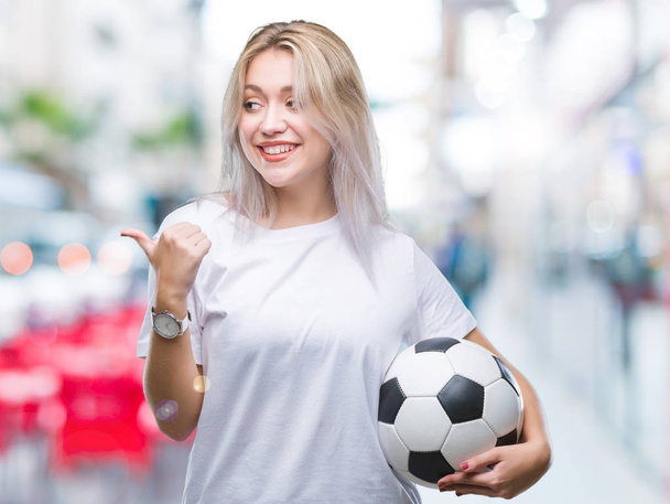 Молода блондинка тримає футбольний м'яч над ізольованим тлом, вказуючи і показуючи з великим пальцем в сторону з щасливим обличчям посміхаючись
 - Фото, зображення