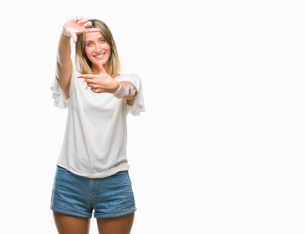 Młoda piękna kobieta na białym tle uśmiechający się podejmowania ramki z rąk i palców z szczęśliwy twarz. Koncepcja kreatywność i fotografii. - Zdjęcie, obraz