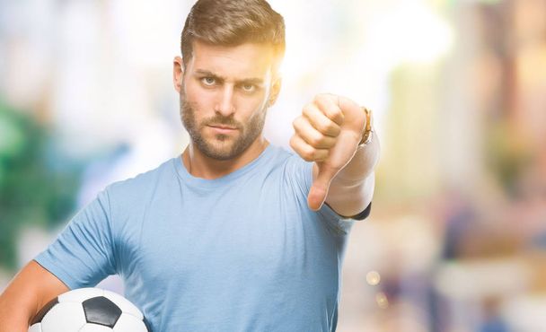 junger gutaussehender Mann hält Fußballball über isoliertem Hintergrund mit wütendem Gesicht, negatives Zeichen zeigt Abneigung mit gesenktem Daumen, Ablehnungskonzept - Foto, Bild