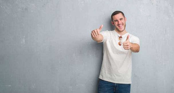 Молодой кавказский мужчина над серой гранж-стеной в солнцезащитных очках, одобряющий позитивный жест рукой, улыбается и радуется успеху. Глядя в камеру, жест победителя
. - Фото, изображение