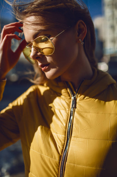 A young European woman, traveling, with long blond hair, wearing a yellow jacket, yellow sunglasses walking down the city center street, street shooting. Even light. - Foto, imagen