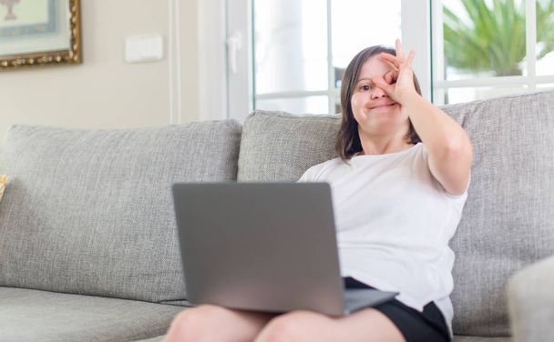 Down syndrome woman at home using computer laptop with happy face smiling doing ok sign with hand on eye looking through fingers - 写真・画像
