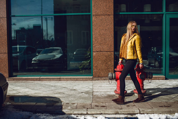 A young European woman, traveling, with long blond hair, wearing a yellow jacket, yellow sunglasses walking down the city center street, street shooting. Even light. - Photo, image