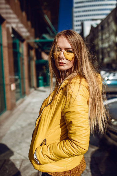 A young European woman, traveling, with long blond hair, wearing a yellow jacket, yellow sunglasses walking down the city center street, street shooting. Even light. - Фото, изображение