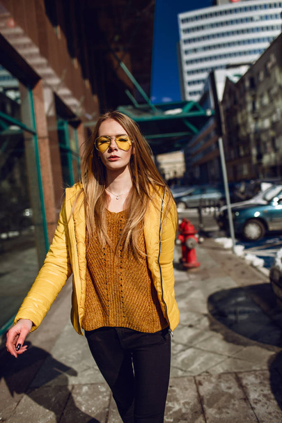 A young European woman, traveling, with long blond hair, wearing a yellow jacket, yellow sunglasses walking down the city center street, street shooting. Even light. - Foto, Bild