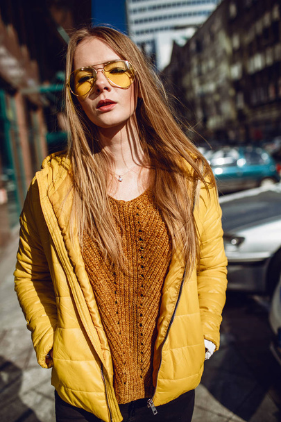 A young European woman, traveling, with long blond hair, wearing a yellow jacket, yellow sunglasses walking down the city center street, street shooting. Even light. - Фото, зображення