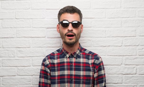 Jonge volwassen man dragen van een zonnebril permanent over witte bakstenen muur bang in schok met een verrassing gezicht, bang en opgewonden met angst expressie - Foto, afbeelding