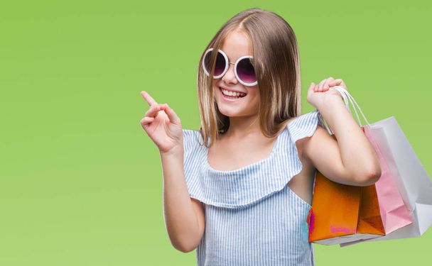 Young beautiful girl holding shopping bags on sales over isolated background very happy pointing with hand and finger to the side - Foto, imagen