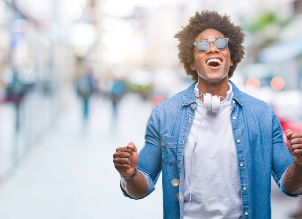 Афро-американец в наушниках, слушающий музыку на изолированном фоне, празднующий безумный и безумный успех с поднятыми руками и с взволнованными закрытыми глазами. Концепция победителя
 - Фото, изображение