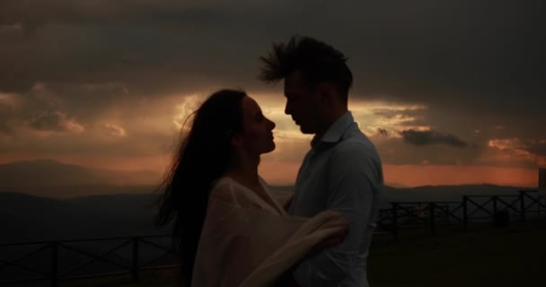 Gone with the wind. Picture of a lovely couple hugging each other tender and kissing while standing on the hill with beautiful red and grey sunset over them - Footage, Video
