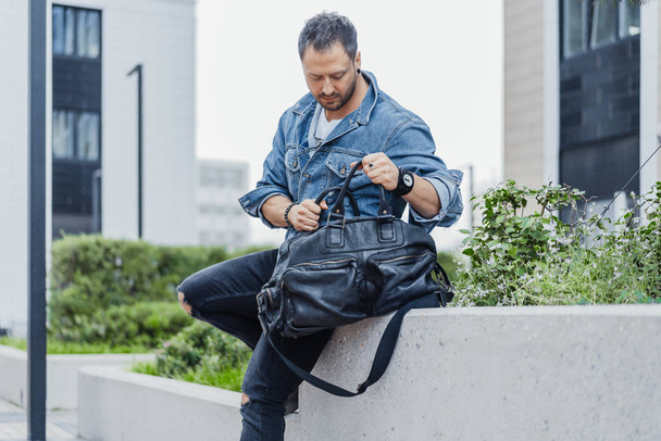 Young man rummaging in black sport bag and sitting on cityscape background. - Fotoğraf, Görsel