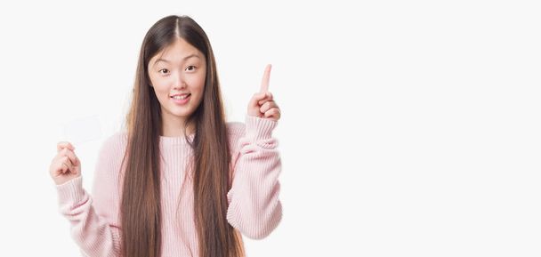 Joven mujer china sobre un fondo aislado sosteniendo la tarjeta de visita sorprendida con una idea o pregunta apuntando con el dedo con la cara feliz, número uno
 - Foto, imagen