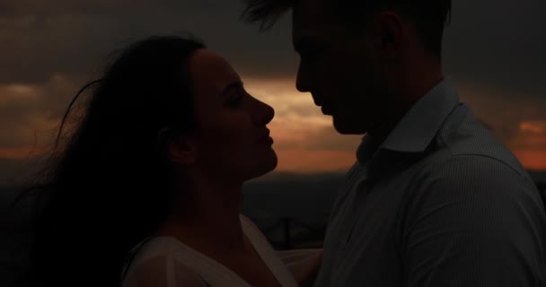 Gegaan met de wind. Foto van een mooie paar knuffelen elkaar mals en zoenen terwijl je op de heuvel met prachtige rode en grijze zonsondergang over hen - Video