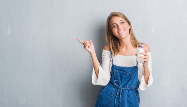 Gyönyörű fiatal nő grunge szürke falon driking egy pohár tej nagyon boldog, mutat, a kéz és az ujjak az oldalon keresztül - Fotó, kép