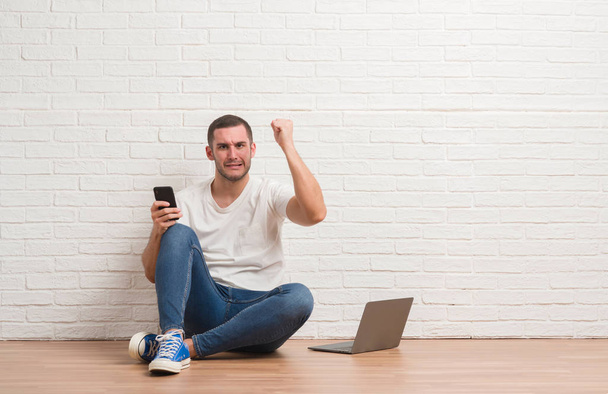 Młody człowiek kaukaski siedzi nad białym murem przy użyciu komputera przenośnego i smartphone zirytowany i sfrustrowany krzycząc z gniewu, szalony i krzyczy ręce, gniew koncepcja - Zdjęcie, obraz
