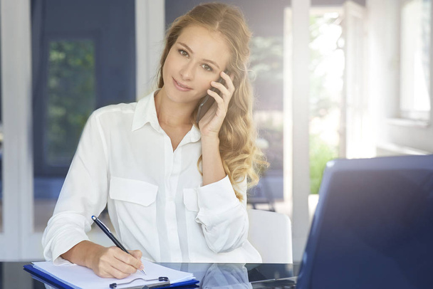 Portrait of beautiful young assistant businesswoman making call while sitting at desk and making notes. - Fotografie, Obrázek