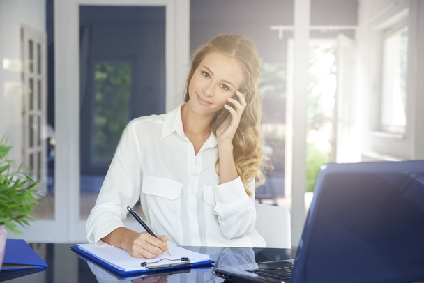 Portrait of beautiful young assistant businesswoman making call while sitting at desk and making notes. - Foto, Imagem