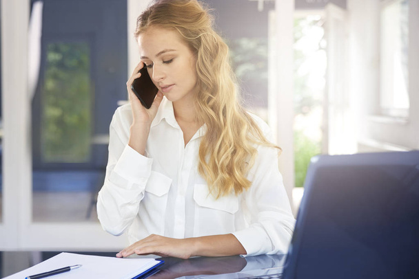 Portrait of beautiful young assistant businesswoman making call while sitting at desk and making notes. - Valokuva, kuva