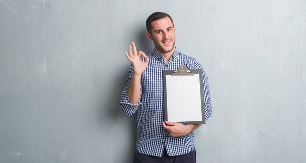 Young caucasian man over grey grunge wall holding clipboard doing ok sign with fingers, excellent symbol - Фото, зображення
