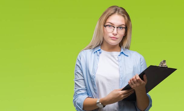 Young caucasian business woman holding clipboard over isolated background with a confident expression on smart face thinking serious - Photo, image