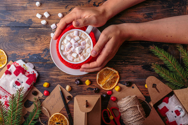 woman hands takes a mug of hot cacao with marshmallows knitted cloth for a mug christmas concept winter background of packing gifts christmas decoration traditional beverage - Fotoğraf, Görsel