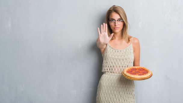 Όμορφη νεαρή γυναίκα πάνω από grunge γκρίζο τοίχο τρώγοντας πίτσα πεπερόνι με ανοιχτό χέρι κάνει στοπ με σοβαρές και αυτοπεποίθηση έκφραση, χειρονομία άμυνα - Φωτογραφία, εικόνα