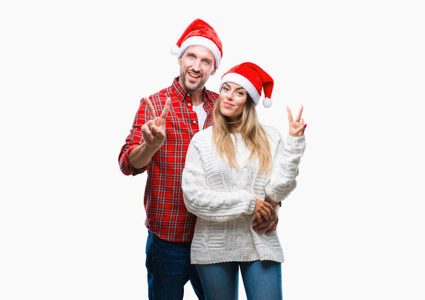 Young couple in love wearing christmas hat over isolated background smiling with happy face winking at the camera doing victory sign. Number two. - Photo, Image