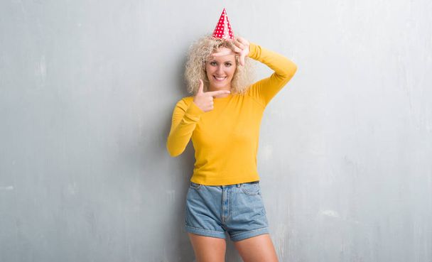 グランジに若いブロンドの女性灰色の壁を手で作るフレームと幸せそうな顔で指を笑顔の誕生日の帽子を身に着けています。創造性と写真のコンセプト. - 写真・画像