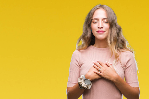 Beautiful young blonde woman over isolated background smiling with hands on chest with closed eyes and grateful gesture on face. Health concept. - Photo, Image