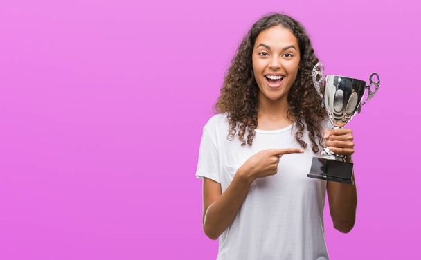 Young hispanic woman holding trophy very happy pointing with hand and finger - Photo, Image