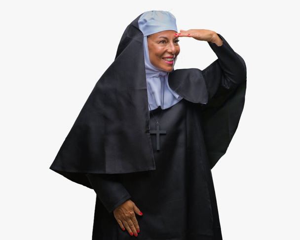 Middelbare leeftijd senior christelijke katholieke non vrouw over geïsoleerde achtergrond erg blij en lachende op zoek ver weg met de hand boven het hoofd. Zoeken op concept. - Foto, afbeelding