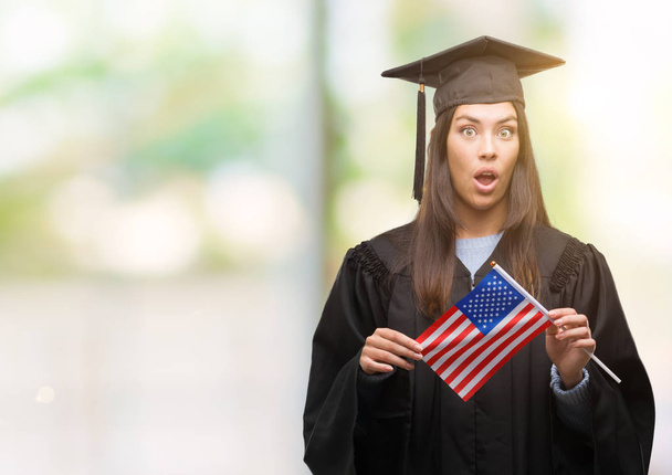 Giovane donna ispanica indossa uniforme graduata tenendo bandiera d'America spaventata in stato di shock con un volto a sorpresa, spaventata ed eccitata con espressione di paura
 - Foto, immagini