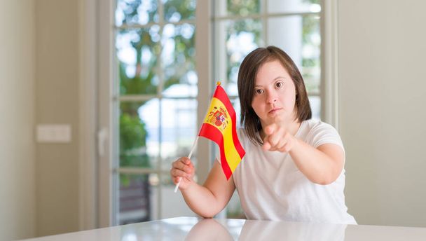 Down-Syndrom-Frau zu Hause mit spanischen Flaggen, die mit dem Finger in die Kamera und zu Ihnen zeigen, Handzeichen, positive und selbstbewusste Geste von vorne - Foto, Bild